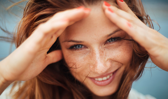 Eine junge FRau mit langen roten Haaren und Sommersprossen lächelt in die Kamera. Sie hält ihre Hände links und rechts an ihr Gesicht, damit der Wind ihr keine Haare ins Gesicht weht.