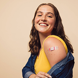 Eine junge Frau mit langen braunen Haaren lacht in die Kamera. Auf ihrem Oberarm klebt ein bunt-gestreiftes Pflaster.