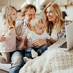 Eine junge Familie sitzt auf dem Sofa. Der Vater und die kleinen Kinder schauen auf einen Laptop. Die Mutter hält den Laptop fest, sie lächelt und schaut ihre Tochter an.