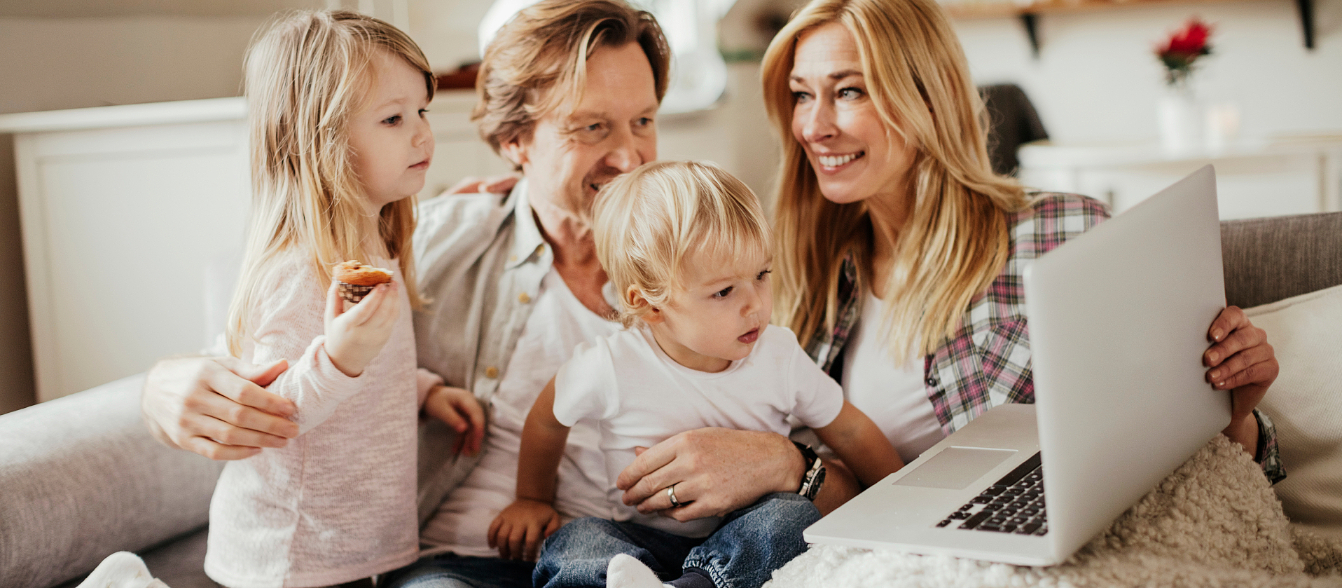 Eine junge vierköpfige Familie liegt zusammen auf dem Sofa. Die Frau umarmt ihren Mann und lacht ihn an.