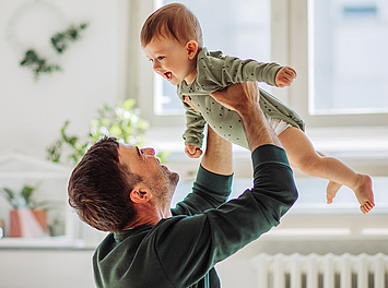 Ein Vater hebt sein Baby in die Luft. Sie lachen beide uns habe Spaß.