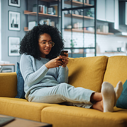 Eine Frau sitzt gemütlich auf einem Sofa und schaut auf ihr Smartphone. 
