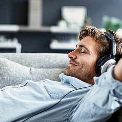 Ein junger Mann liegt mit geschlossenen Augen auf dem Sofa, lächelt zufrieden und trägt dabei Kopfhörer.