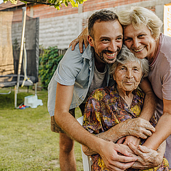 Ein Mann und eine Frau umarmen gemeinsam eine Seniorin. Sie sind dabei im Garten.