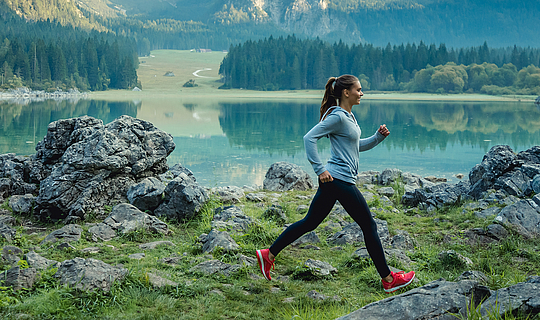 Eine junge Frau mit langen braunen Haaren joggt an einem See. Sie trägt Sportkleidung und knallrote Turnschuhe. Der Untergrund ist sehr felsig, im Hintergrund sieht man einen Wald und Berge. 