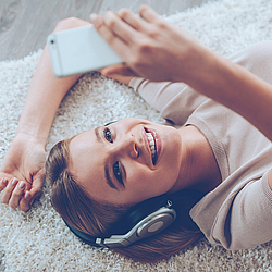 Eine Frau mit braunen Haaren liegt auf einem flauschigen Teppich. Sie trägt silberne Kopfhörer und hält ihr Handy in der Hand.