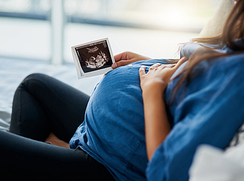Eine schwangere Frau sitzt auf dem Sofa. In der rechten Hand hält sie ein Ultraschallbild ihres Babys.