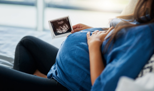 Eine junge schwangere Frau liegt auf dem Sofa. Ihre linke Hand liegt auf ihrem Bauch, in der rechten Hand hält sie ein Ultraschallbild ihres Babys.