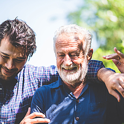 Ein junger Mann mit dunklem Haar und Bart legt seinen Arm um seinen weißhaarigen Vater. Beide lachen.