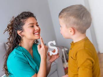 Eine junge Logopädin mit langen lockigen Haaren zeigt auf ihren Mund. Sie übt mit einem kleinen blonden Jungen die Aussprache des Buchstabens S.