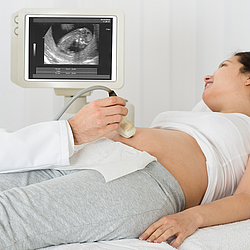 Eine junge schwangere Frau liegt auf einer Untersuchungsliege. Ein Arzt macht einen Ultraschall von ihrem Bauch.