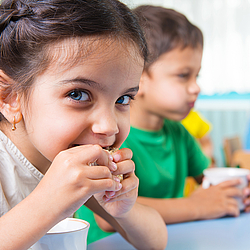 Ein kleines Mädchen mit langen braunen Haaren schaut fröhlich in die Kamera und beißt in ein Brot.