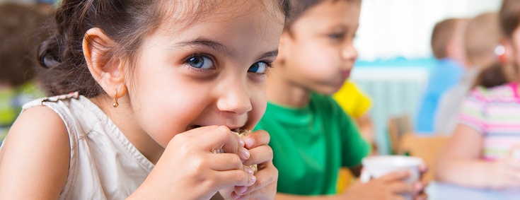 Ein kleines Mädchen mit langen braunen Haaren schaut fröhlich in die Kamera und beißt in ein Brot. Im Hintergrund sieht man noch weitere Kinder beim Essen und Trinken.