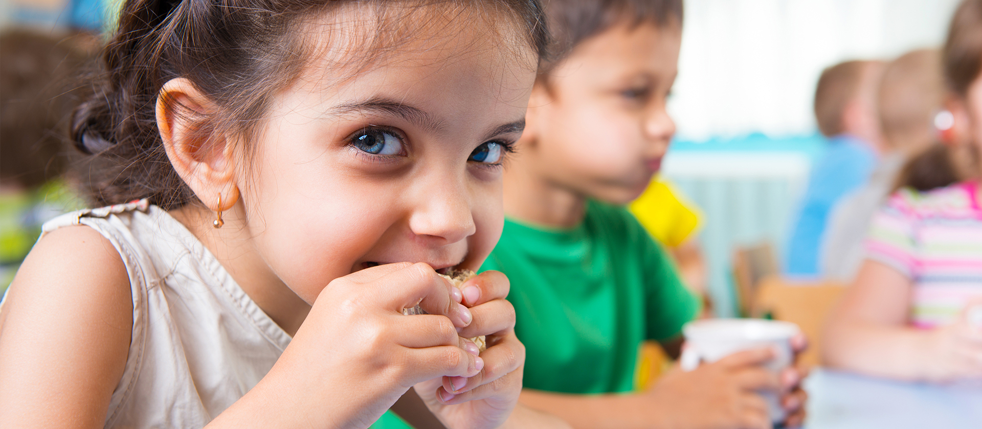 Ein kleines Mädchen mit langen braunen Haaren schaut fröhlich in die Kamera und beißt in ein Brot. Im Hintergrund sieht man noch weitere Kinder beim Essen und Trinken.