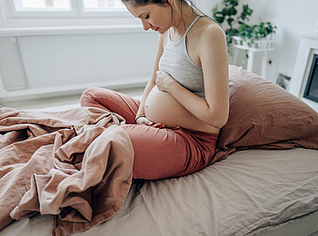 Eine junge schwangere Frau sitzt in ihrem Bett. Sie hat ihre Hände an ihren Bauch gelegt und lächelt.