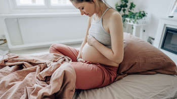 Eine junge schwangere Frau sitzt in ihrem Bett. Sie hat ihre Hände oben und unten an ihren Bauch gelegt und schaut lächelnd auf ihren Bauch herunter. 
