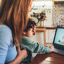 Eine Frau sitzt mit ihrem Kind am Tisch. Die Frau hält dem Kind die Stirn. Sie schauen auf einen Laptop mit einem Online-Arzt.