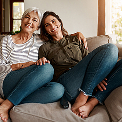 Eine ältere Frau mit kurzen grauen Haaren sitzt neben ihrer Tochter auf dem Sofa. Beide lachen fröhlich in die Kamera.