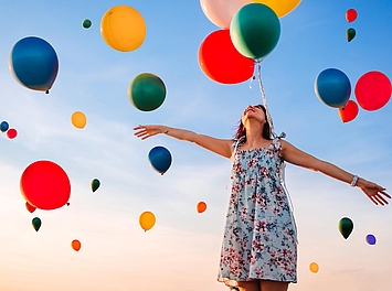 Person mit ausgestreckten Armen unter bunten Luftballons vor blauem Himmel.
