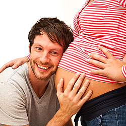 Ein junger Mann hält lächelnd sein Ohr an den Bauch seiner schwangeren Partnerin, eine Hand liegt auf ihrem Bauch. 