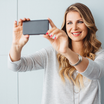 Eine junge Frau mit langen blonden Haaren steht vor einer grauen Wand und lächelt. Sie macht ein Selfie mit ihrem Handy.