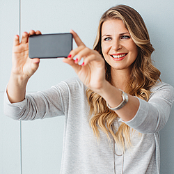 Eine Frau mit langen blonden Haaren steht vor einer grauen Wand und lächelt. Sie macht ein Selfie mit ihrem Handy.