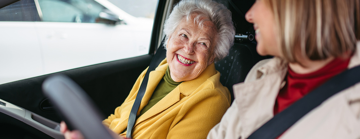 Eine ältere Frau mir kurzen grauen Haaren trägt einen gelben Mantel. Sie sitzt in einem Auto auf dem Beifahrersitz. Neben ihr sitzt eine junge Frau mit schulterlangen hellen Haaren und einem beigefarbenen Mantel. Die beiden schauen sich lächelnd an.