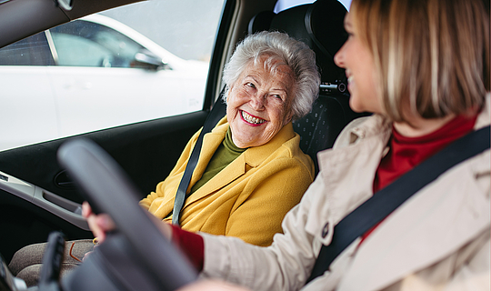 Eine ältere Frau mir kurzen grauen Haaren und eine junge Frau mit schulterlangen hellen Haaren sitzen in Auto und schauen sich an.