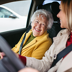 Eine ältere Frau mir kurzen grauen Haaren und eine junge Frau mit schulterlangen hellen Haaren sitzen in Auto und schauen sich an.