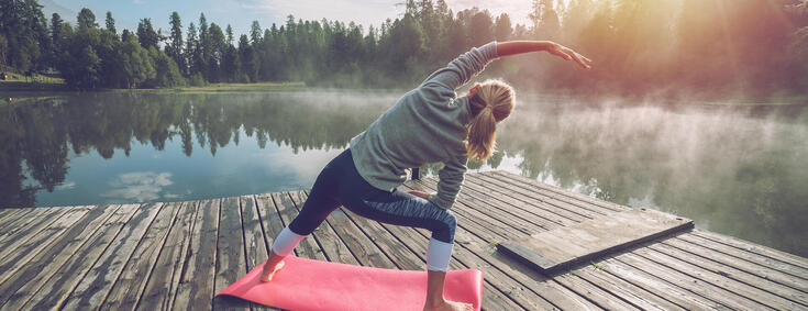 Frau auf einem Steg am Wasser macht Yoga-Übungen in der Natur