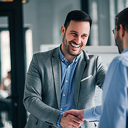 Ein Mann mit Anzug schüttelt einem anderen Mann mit Brille die Hand. Er lacht fröhlich.