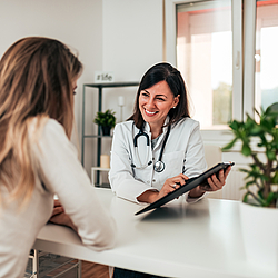 Eine Ärztin sitzt gegenüber einer Patientin. Sie erklärt ihr etwas und zeigt dabei auf ein Klemmbrett mit Unterlagen.  