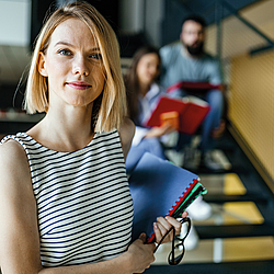 Eine Frau mit kurzen blonden Haare schaut lächelnd in die Kamera. In der Hand hält sie Hefter und eine Brille. 