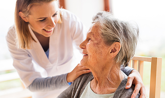 Eine Pflegekraft mit schulterlangen blonden Haaren hat ihre Hände auf die Schultern einer älteren Patientin gelegt. Sie lächelt die Patientin an.