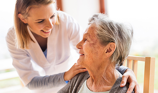 Eine junge Pflegekraft mit schulterlangen blonden Haaren hat ihre Hände auf die Schultern einer älteren Patientin gelegt. Sie lächelt die Patientin aufmunternd an.