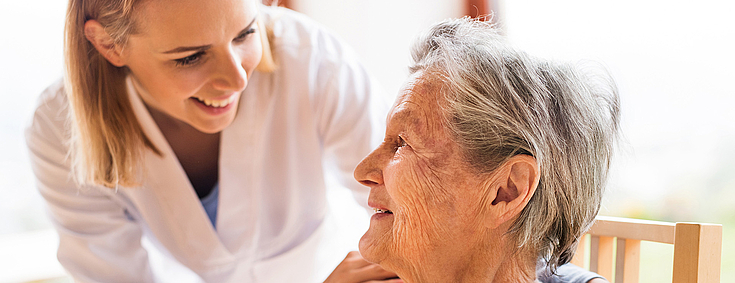 Eine junge Pflegekraft mit schulterlangen blonden Haaren hat ihre Hände auf die Schultern einer älteren Patientin gelegt. Sie lächelt die Patientin aufmunternd an.