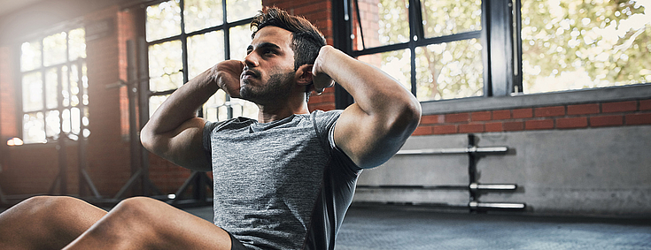 Ein junger Mann mit dunklen Haaren ist im Fitnessstudio. Er trägt kurze Sportkleidung und macht Übungen auf einer blauen Sportmatte.