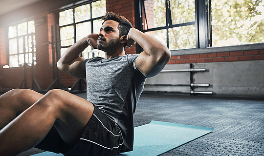 Ein junger Mann mit dunklen Haaren ist im Fitnessstudio. Er trägt kurze Sportkleidung und macht Übungen auf einer blauen Sportmatte.