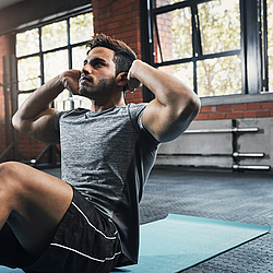 Ein junger Mann mit dunklen Haaren ist im Fitnessstudio. Er trägt kurze Sportkleidung und macht Übungen auf einer blauen Sportmatte.