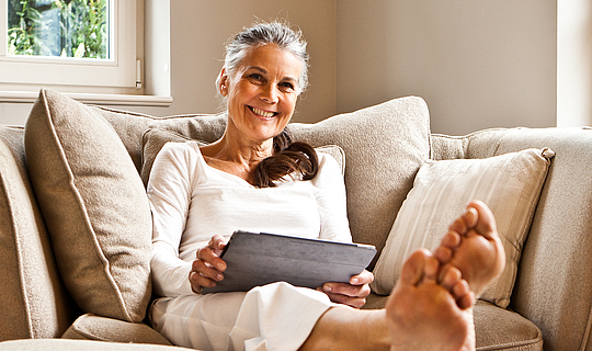Eine Frau mit langen grauen Haare sitzt auf dem Sofa, sie hat die Füße hochgelegt. Sie trägt weiße Kleidung, hält ein Tablet in den Händen und lacht fröhlich in die Kamera.