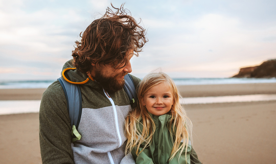 Ein Mann mit langen lockigen Haaren schaut lächelnd auf seine Tochter. Das Mädchen hat lange blonde Haare und lächelt in die Kamera. Die beiden sind an einem Strand.