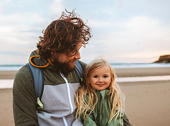 Ein Mann mit langen lockigen Haaren schaut lächelnd auf seine Tochter. Die beiden sind an einem Strand.
