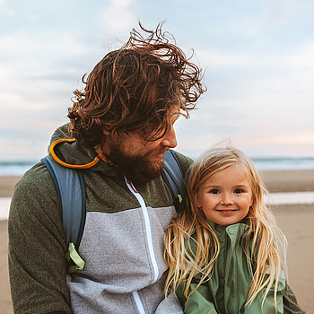 Ein Mann mit langen lockigen Haaren schaut lächelnd auf seine Tochter. Das Mädchen hat lange blonde Haare und lächelt in die Kamera. Die beiden sind an einem Strand.