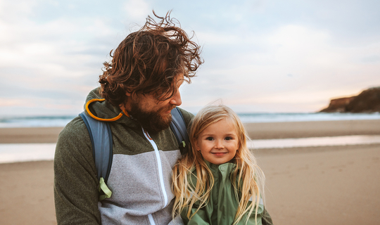 Ein Mann mit langen lockigen Haaren schaut lächelnd auf seine Tochter. Das Mädchen hat lange blonde Haare und lächelt in die Kamera. Die beiden sind an einem Strand.