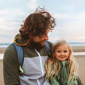 Ein Mann mit langen lockigen Haaren schaut lächelnd auf seine Tochter. Das Mädchen hat lange blonde Haare und lächelt in die Kamera. Die beiden sind an einem Strand.