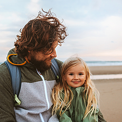 Ein Mann mit langen lockigen Haaren schaut lächelnd auf seine Tochter. Die beiden sind an einem Strand.