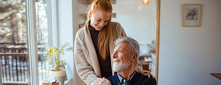 Eine Frau legt einem älteren Mann im Rollstuhl ihre Hände auf die Schultern.