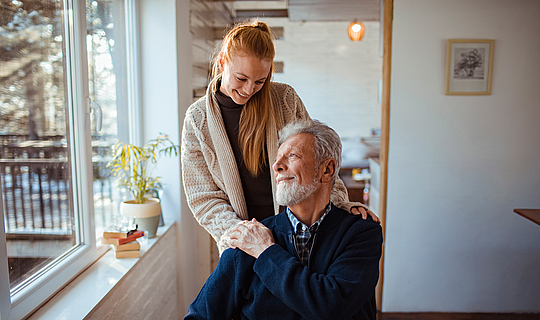 Eine Frau legt einem älteren Mann im Rollstuhl ihre Hände auf die Schultern.