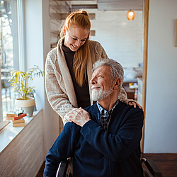 Eine Frau legt einem älteren Mann im Rollstuhl ihre Hände auf die Schultern.