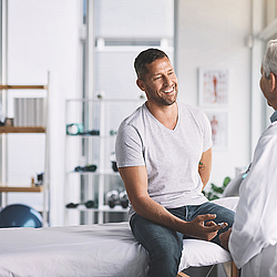 Ein Mann sitzt auf einer Behandlungsliege und lächelt seinen Arzt an. Der Arzt im weißen Kittel lächelt zurück.